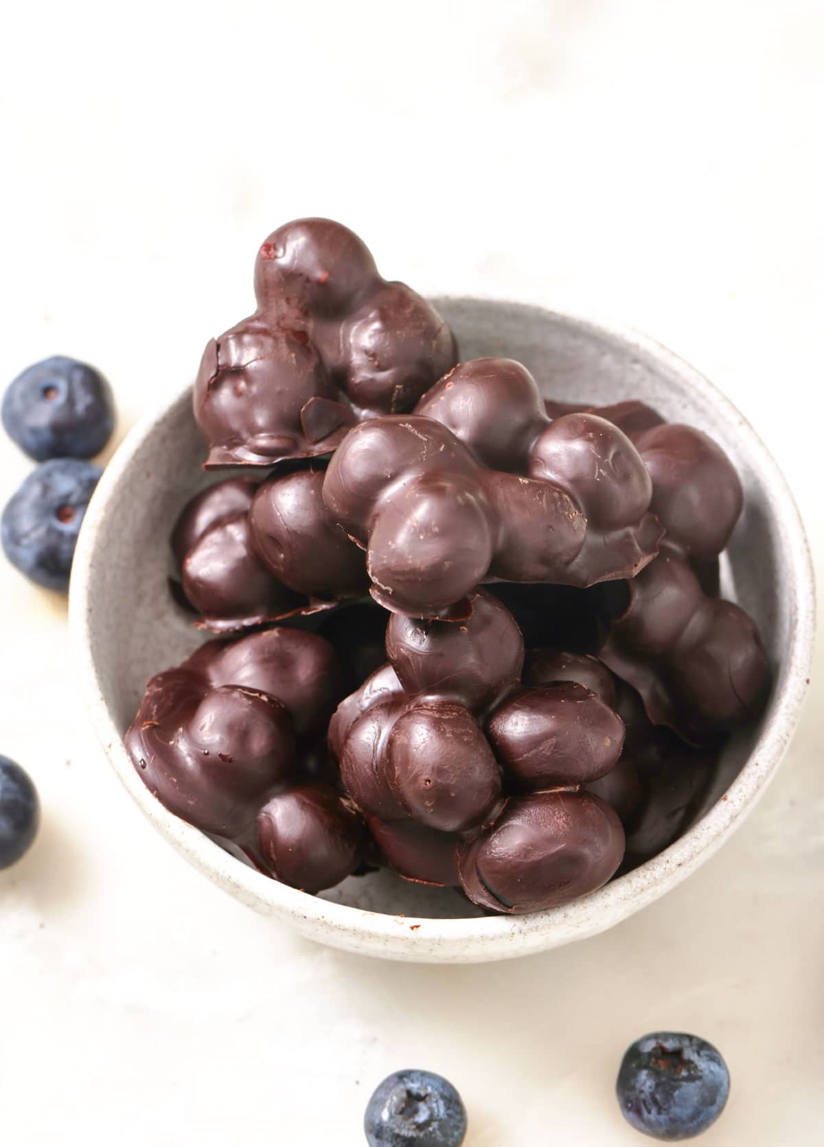 A bowl with chocolate covered blueberry clusters surrounded by fresh blueberries.