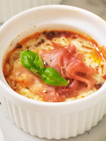 A ramekin with cottage cheese pizza bowl topped with melted cheese, ham and basil leaf.