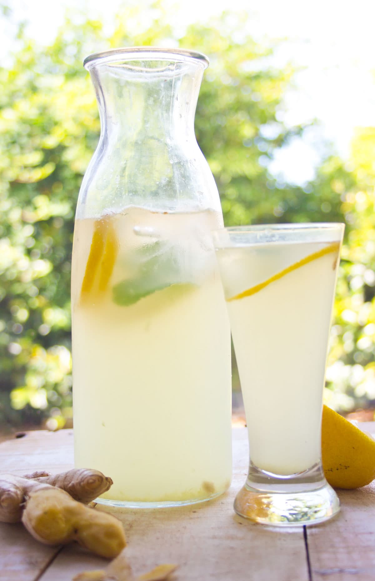 A jug with sugar free ginger ale and a filled glass surrounded by half a lemon and a ginger root.