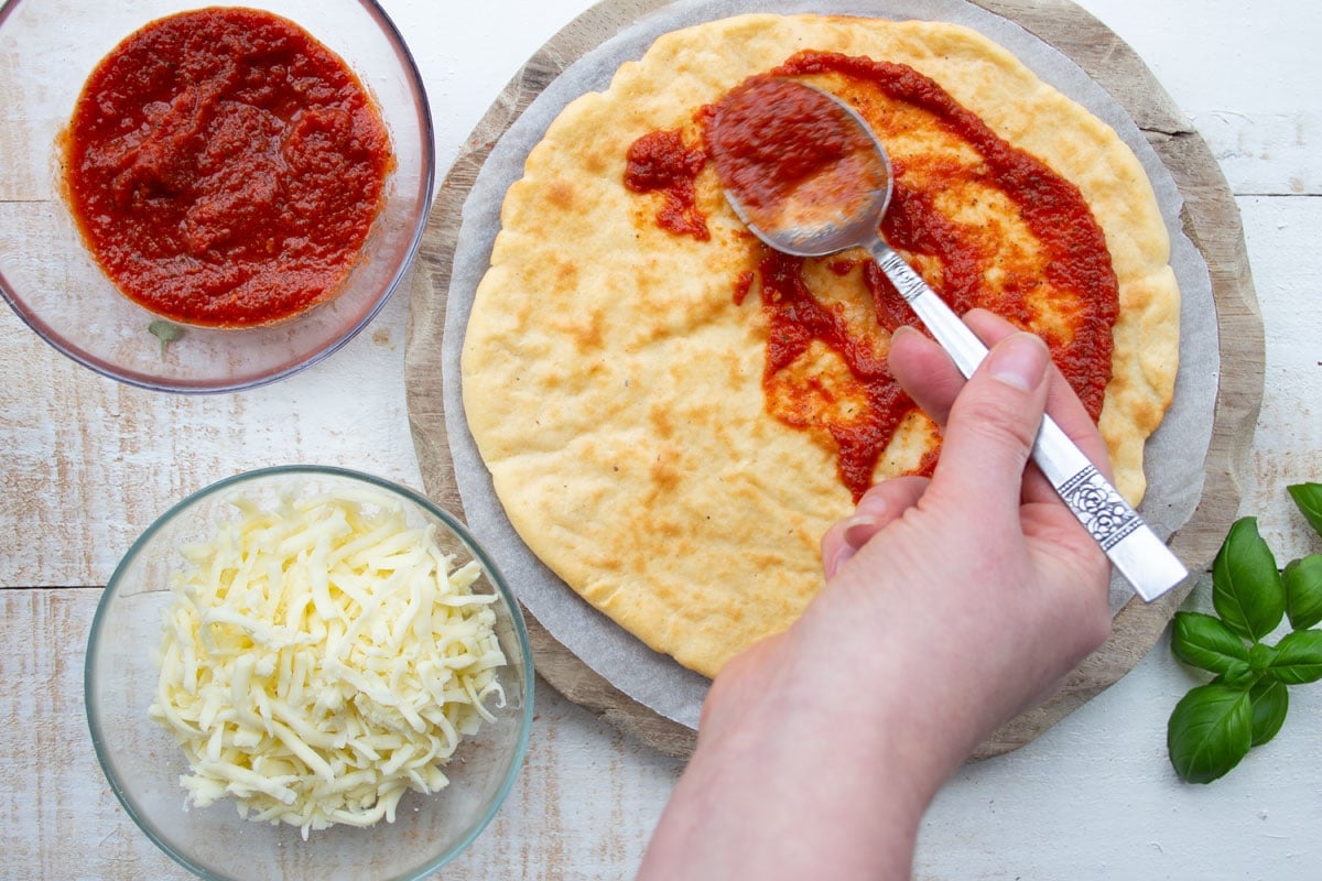 Spreading the baked pizza base with marinara sauce using a spoon.