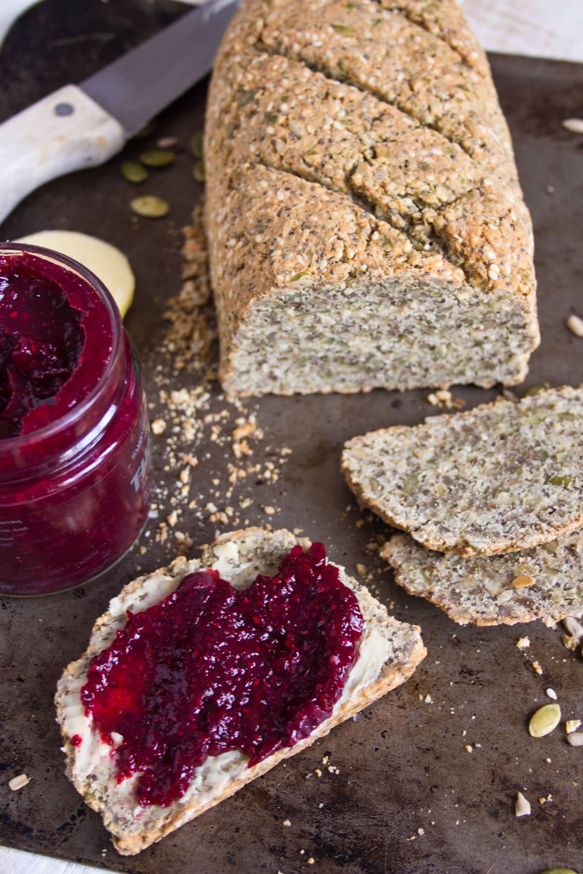 A loaf of seeded bread and a slice of bread that is buttered and spread with jam.