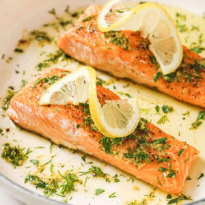 2 keto salmon fillets on a plate topped with olive oil herb dressing and lemon slices.