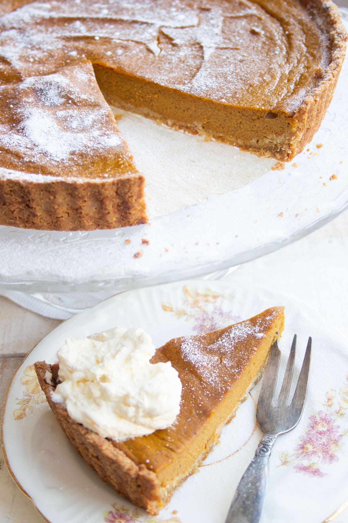 A slice of pumpkin pie topped with whipped cream on a plate with a fork and the pumpkin pie on a cake stand.