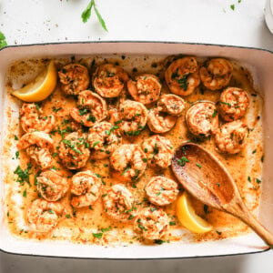 Oven baked shrimp in a rectangular tray topped with chopped parsley and decorated with lemon wedges and a serving spoon.