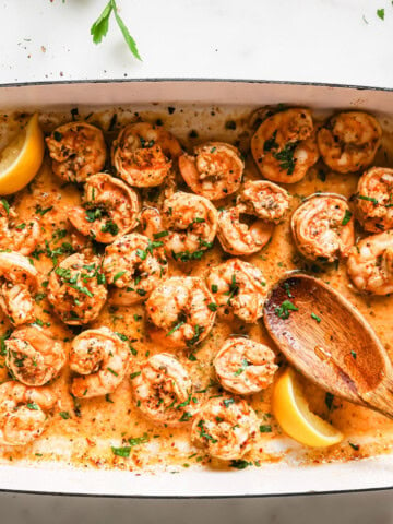 Oven baked shrimp in a rectangular tray topped with chopped parsley and decorated with lemon wedges and a serving spoon.