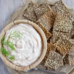 A wooden board with flax and sesame crackers and a bowl with taramasalata dip.