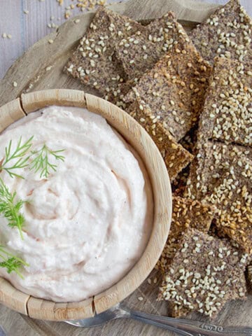 A wooden board with flax and sesame crackers and a bowl with taramasalata dip.