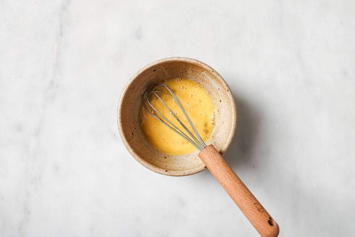 Beaten egg in a bowl and a balloon whisk.