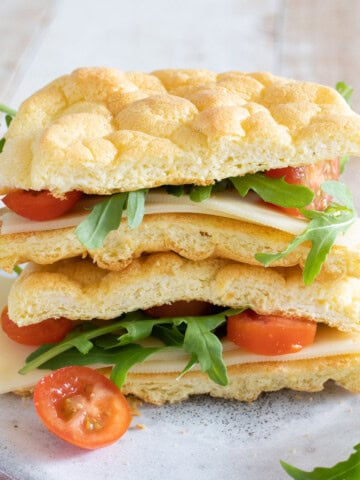 A sandwich made with cloud bread and filled with cheese and lettuce and tomato sliced, sliced open in half.