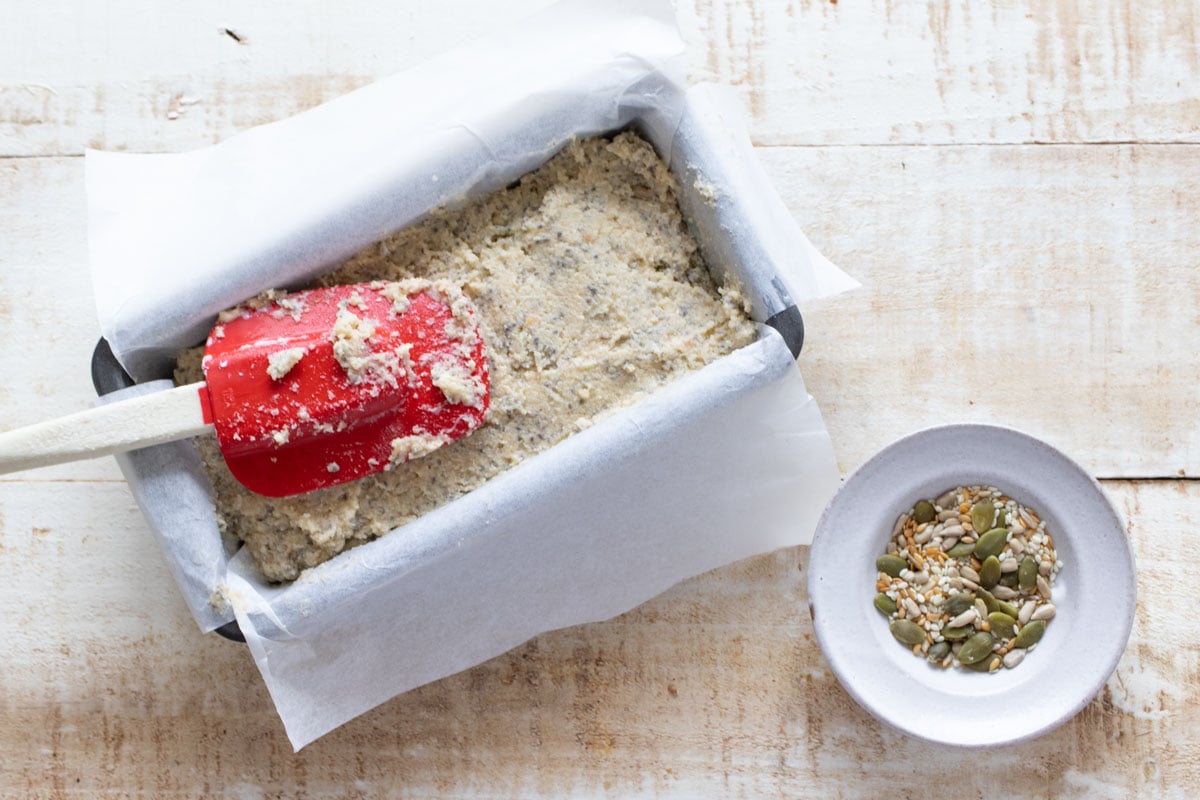 Spreading the dough into the bread pan lined with parchment paper and a bowl with mixed seeds.