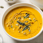 A bowl with keto pumpkin soup topped with pumpkin seeds, olive oil drizzle and fried sage leaves.