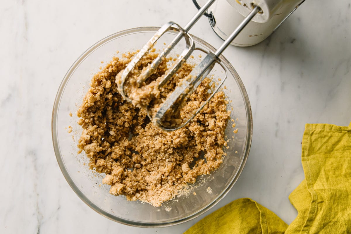A bowl with crumbly cookie dough made from nut flour and melted butter and an electric mixer.