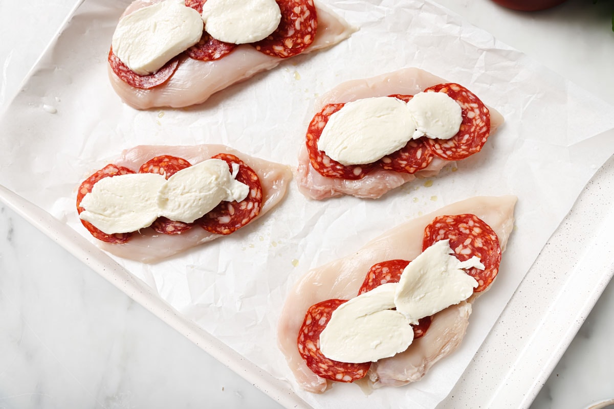 Chicken fillets in a casserole on parchment paper topped with salami and mozzarella slices.