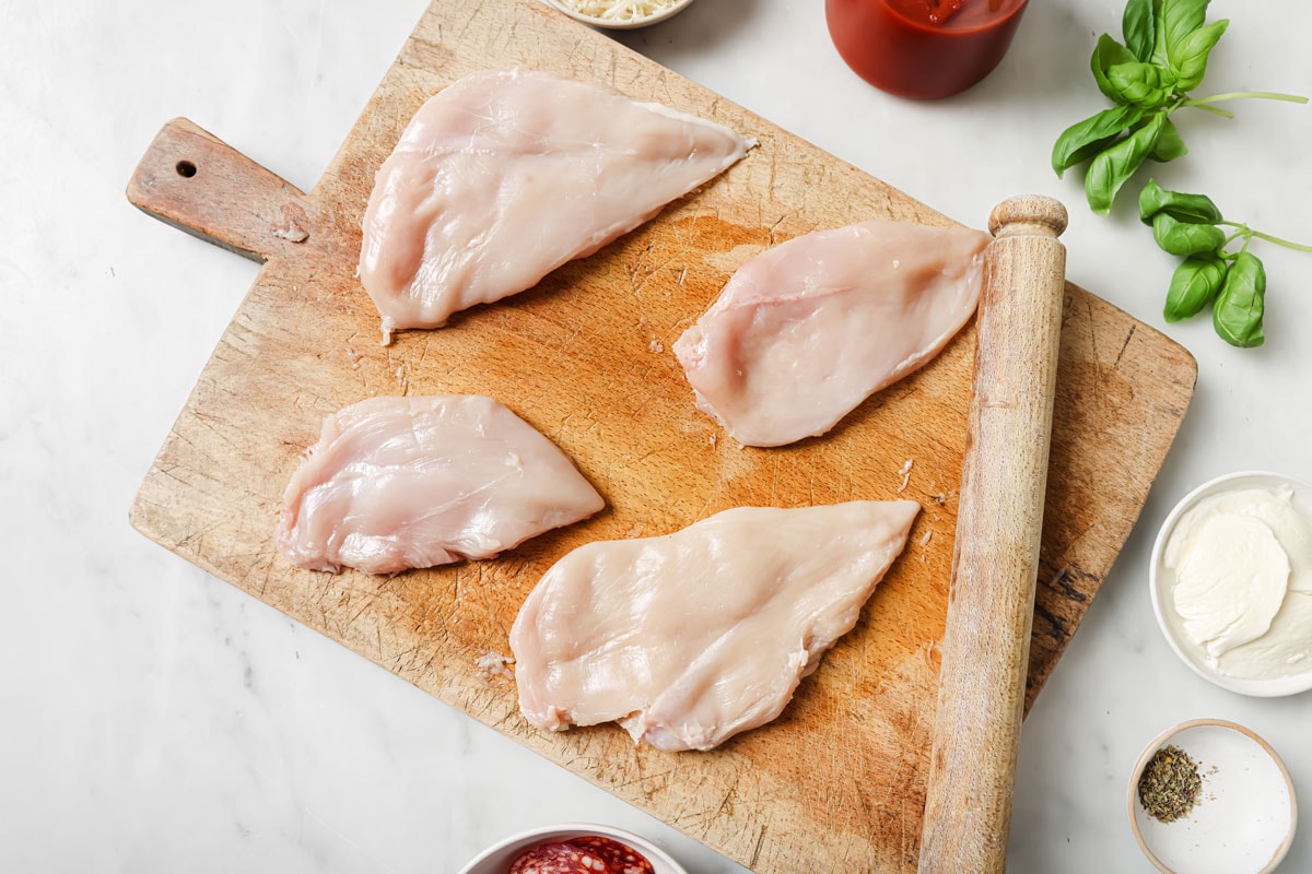 Flattened chicken fillets on a wooden board and a rolling pin.