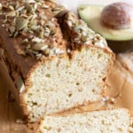 A loaf of avocado bread sliced open and topped with pumpkin seeds and an avocado half in the background.