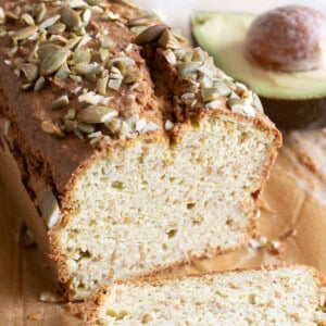 A loaf of avocado bread sliced open and topped with pumpkin seeds and an avocado half in the background.