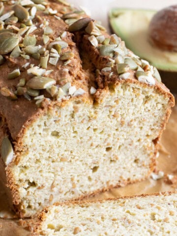 A loaf of avocado bread sliced open and topped with pumpkin seeds and an avocado half in the background.