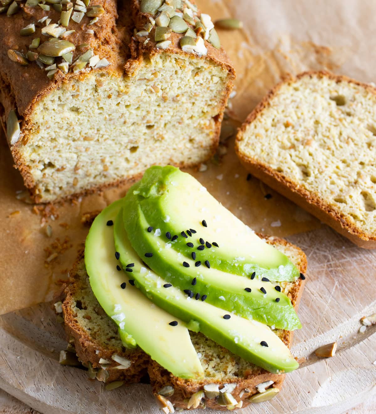 A slice of avocado bread topped with sliced avocado pieces plus salt and nigella seeds and a loaf of avocado bread.