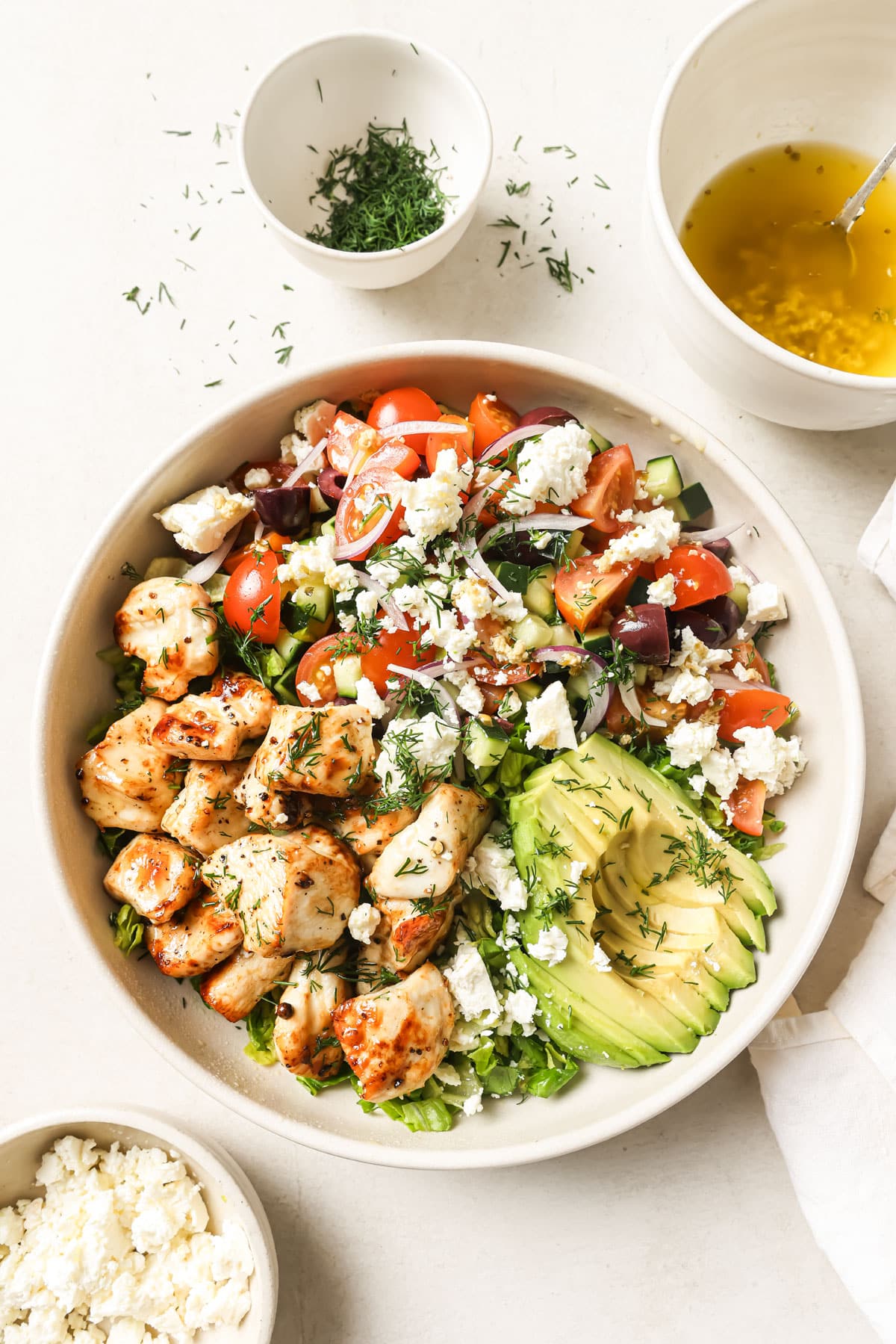 Assembling the Mediterranean chicken salad in a large bowl with the chopped lettuce at the bottom topped with sliced avocado, the seared chicken cubes and the Greek salad, finished off with fresh herbs.