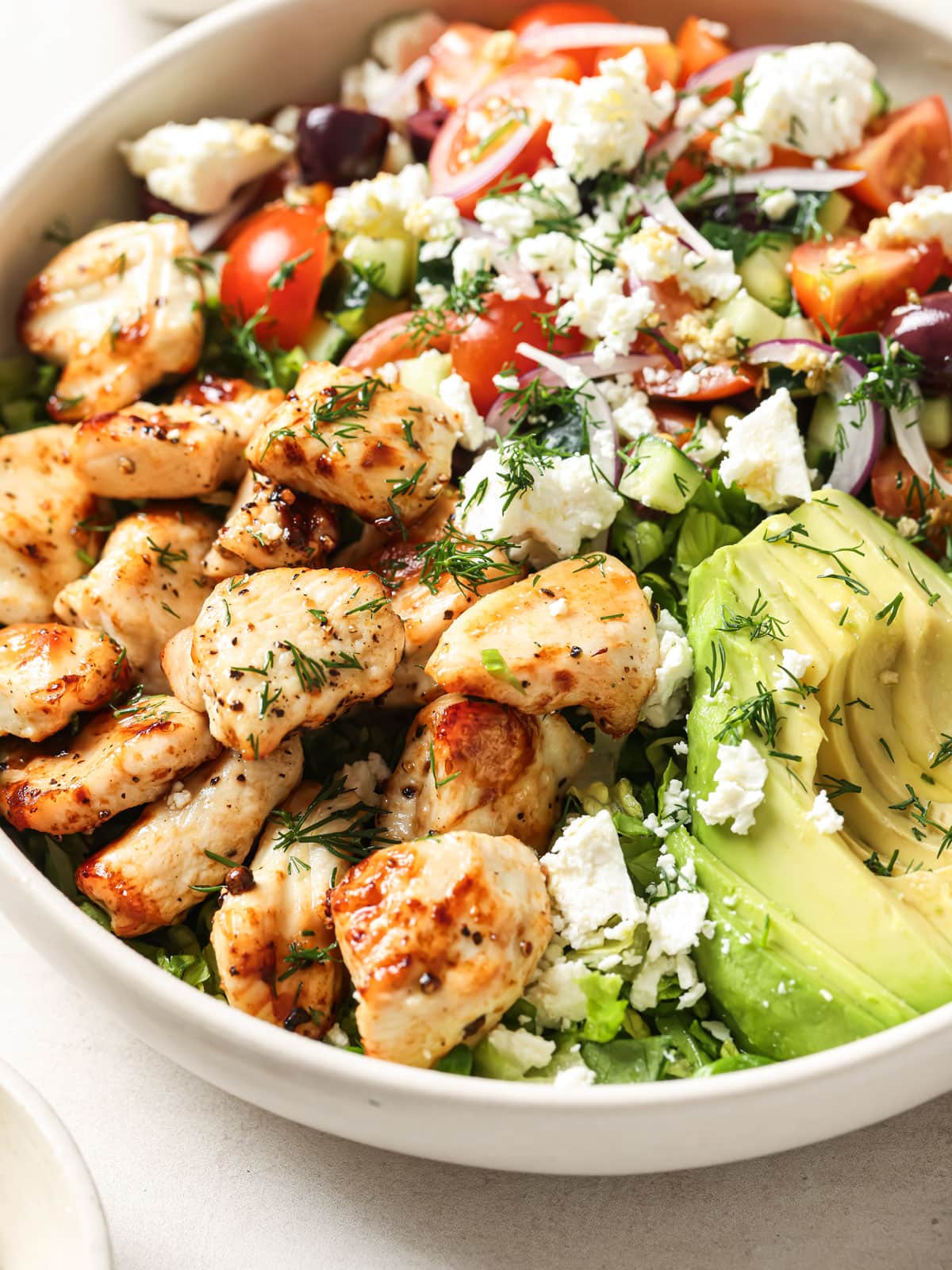 A mediterranean chicken salad with seared chicken breast cubes, feta, avocado, cucumber, lettuce and herbs in a bowl.