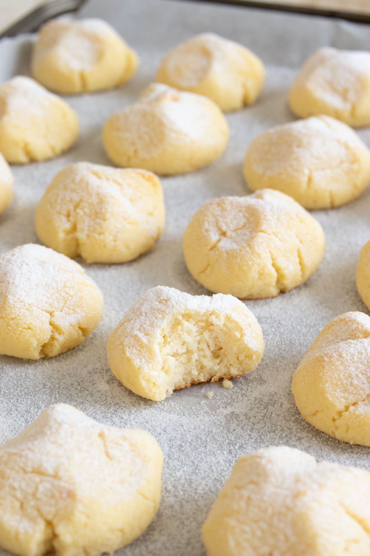 Cottage cheese cookies dusted with powdered sweetener on a baking sheet lined with parchment paper, one is bitten into.