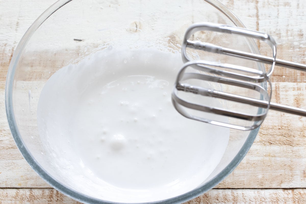 A mixing bowl with whippe egg white and sweetener and two metal mixer attachments being lifted out from it.
