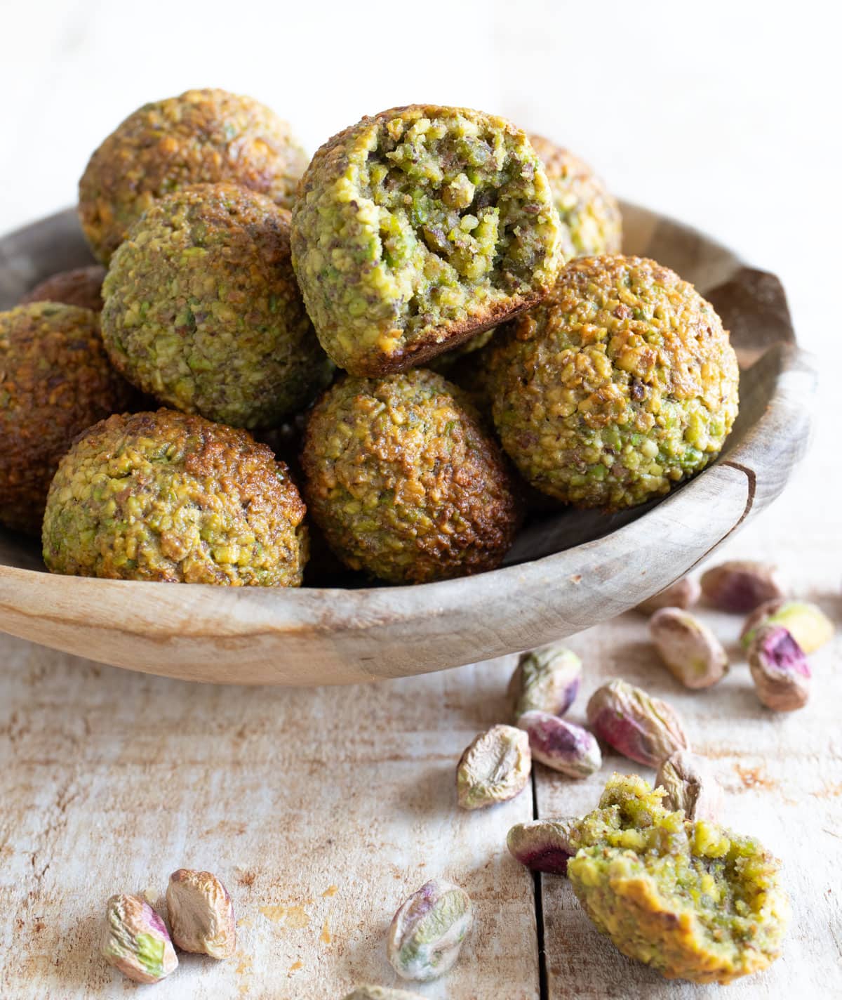 Low carb pistachio cookies in a wooden bowl, one has a piece broken off. This is lying outside the bowl with pistachios.