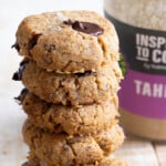 A stack of keto tahini cookies with chocolate chips and topped with sea salt flakes, with a jar of tahini in the background.