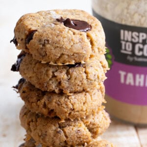 A stack of keto tahini cookies with chocolate chips and topped with sea salt flakes, with a jar of tahini in the background.