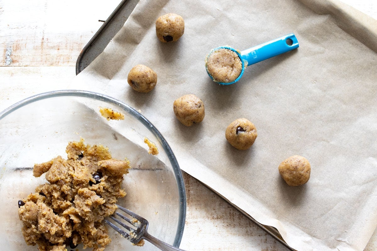 Scooping cookie dough with a tablespoon onto a parchment paper lined baking sheet.
