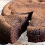 A slice of cottage cheese chocolate cake dusted with cocoa powder being taken from a cake on a cake stand.