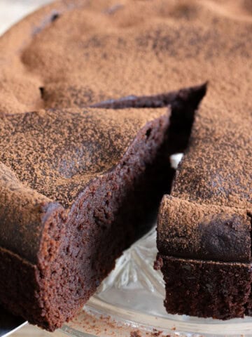 A slice of cottage cheese chocolate cake dusted with cocoa powder being taken from a cake on a cake stand.