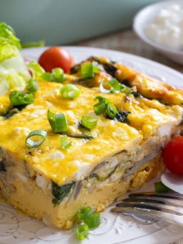 A slice of cottage cheese egg bake with sounach and musnrooms topped with spring onion on a plate with cherry tomatoes and chopped letuce.