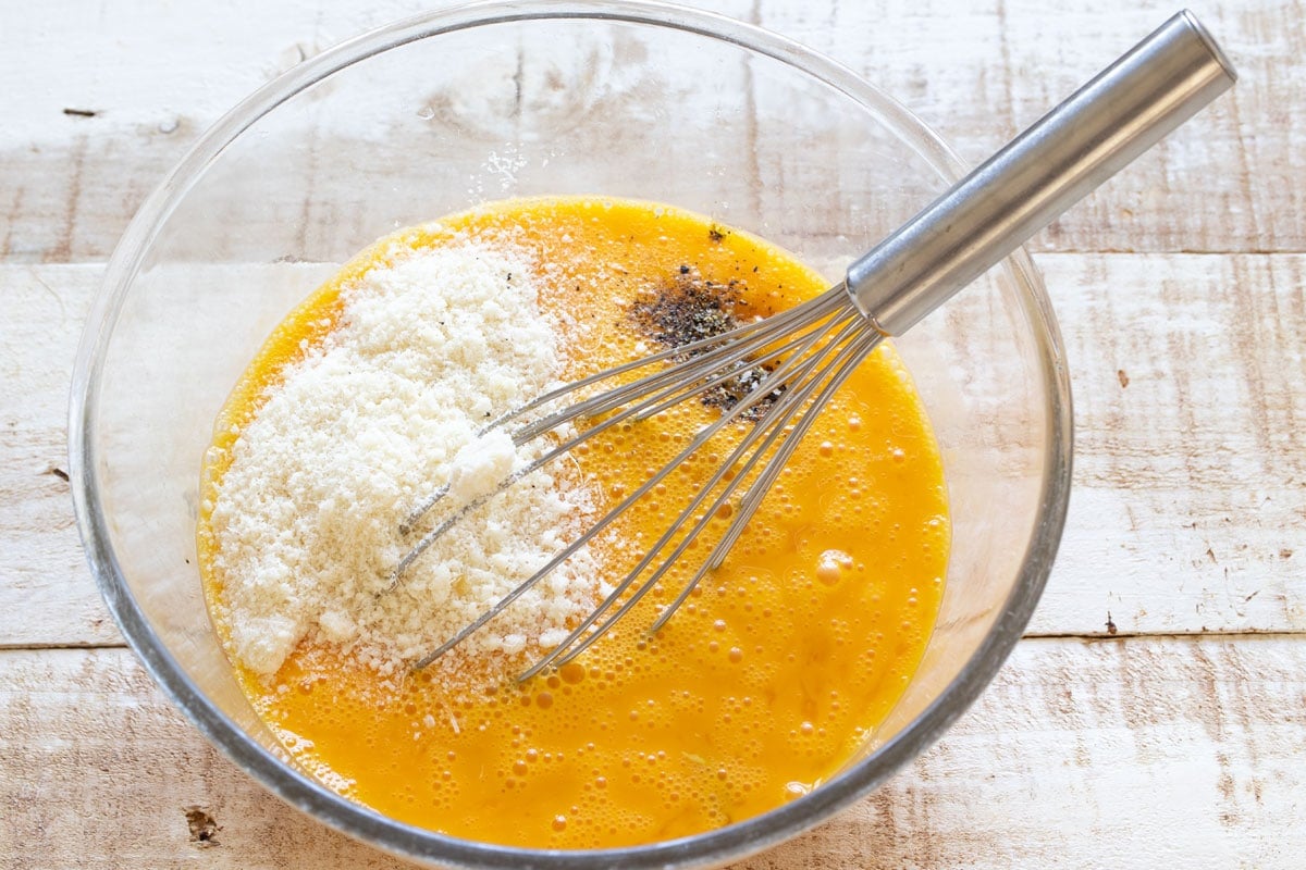 Grated parmesan cheese and seasonings with beaten eggs in a bowl with a balloon whisk.