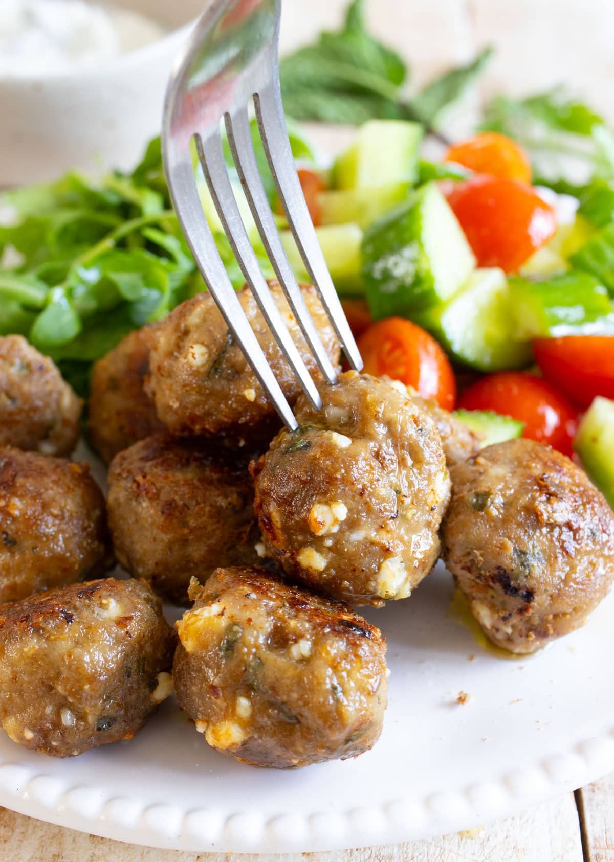 Closeup of a fork picking up a greek lamb meatball from a plate with meatballs and a mixed salad.
